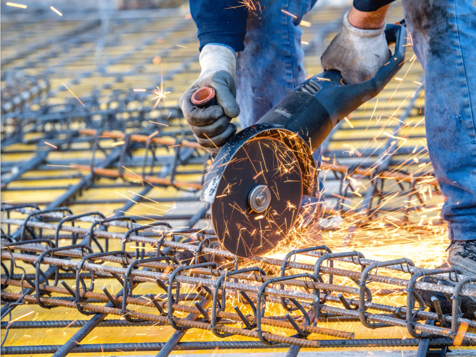 Construction worker using an angle grinder to cut steel rebar with sparks flying at a job site, illustrating factors that can impact overall construction permit cost.