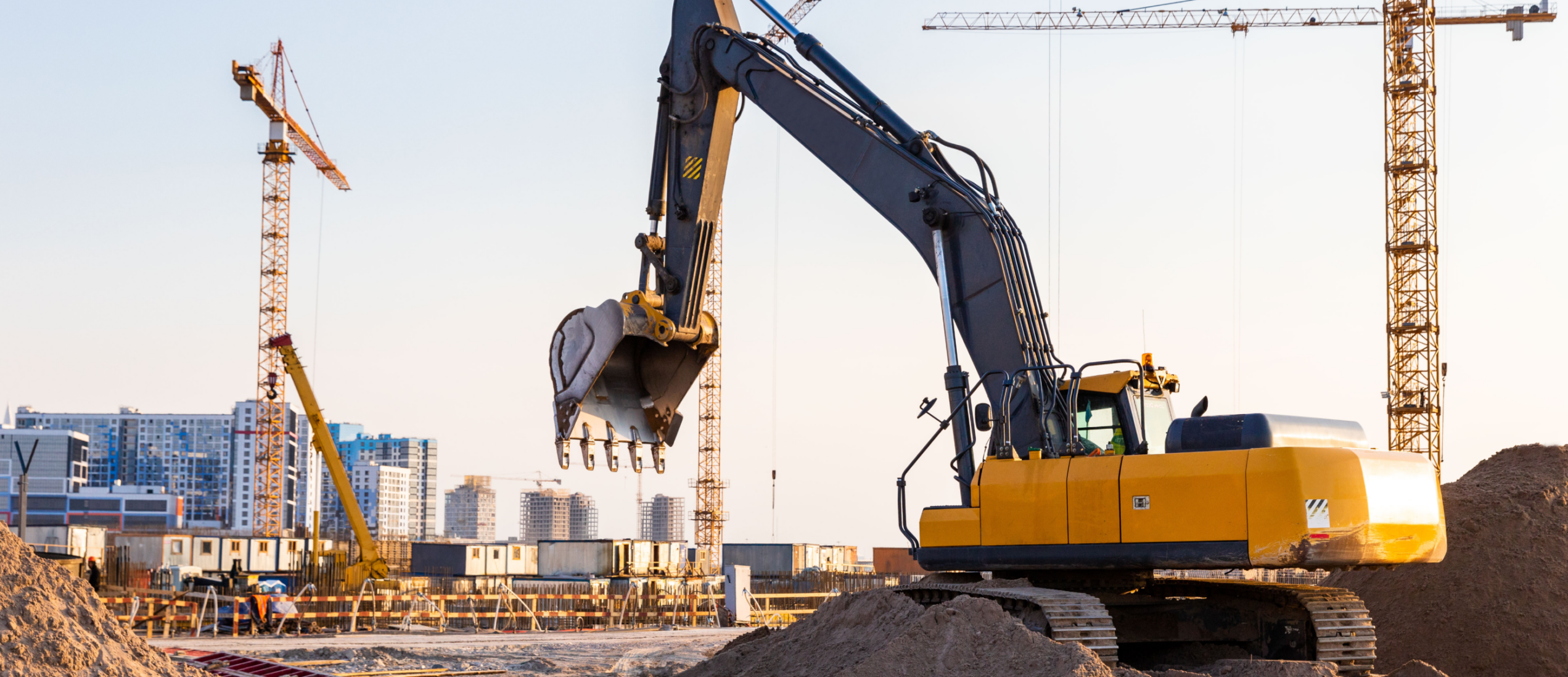 Excavator operating on a large construction site with cranes and partially built structures in the background, illustrating construction safety challenges in complex, active work environments