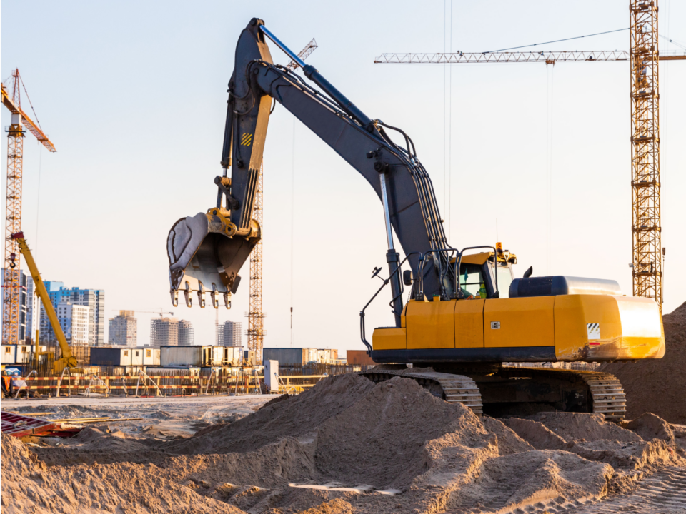Excavator operating on a large construction site with cranes and partially built structures in the background, illustrating construction safety challenges in complex, active work environments